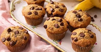 Freshly baked banana chocolate chip muffins arranged on a serving platter with ripe bananas in the background.